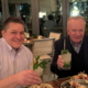 two men sitting at a table with food and drinks