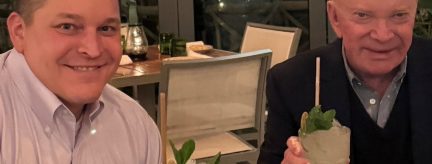 two men sitting at a table with food and drinks